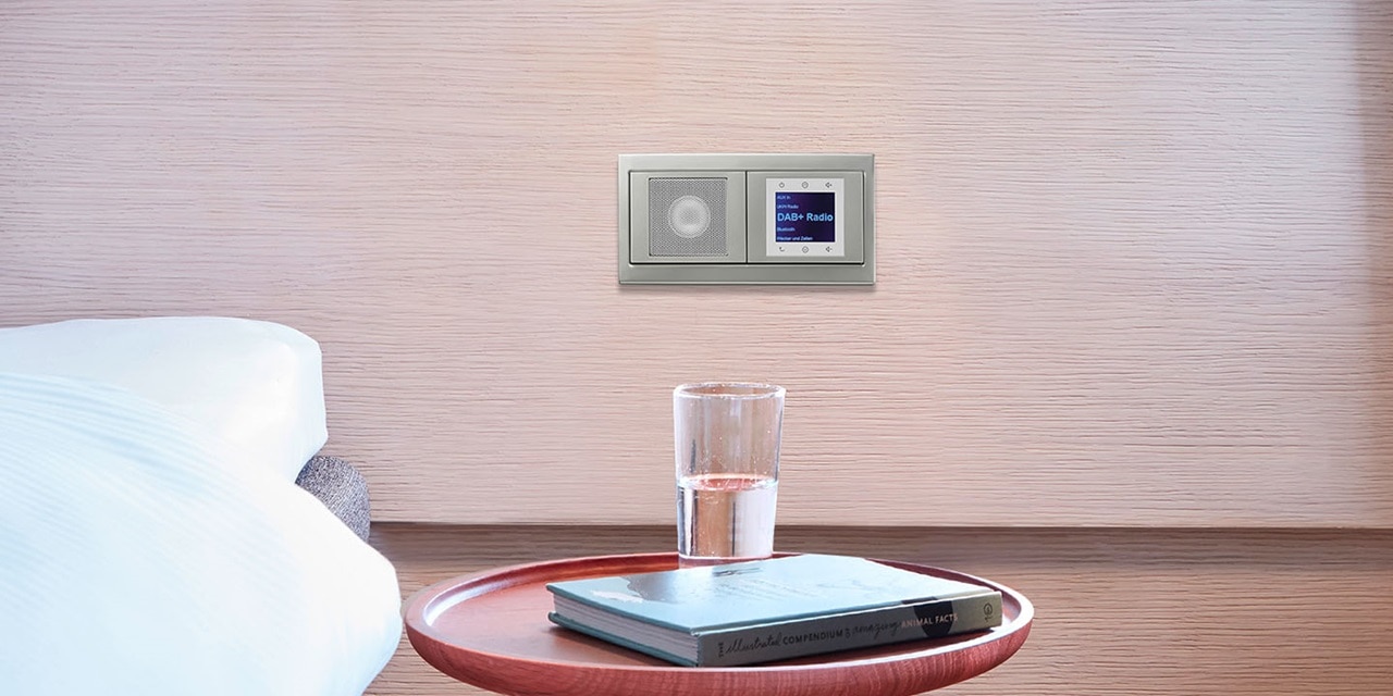 An der Wand montiertes Smart-Home-Panel mit Touchscreen und Lautsprecher neben einem Nachttisch mit einem Glas Wasser und Büchern