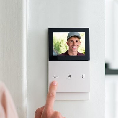 Person benutzt eine wandmontierte Gegensprechanlage mit Videoanzeige, auf der eine lächelnde Person zu sehen ist
