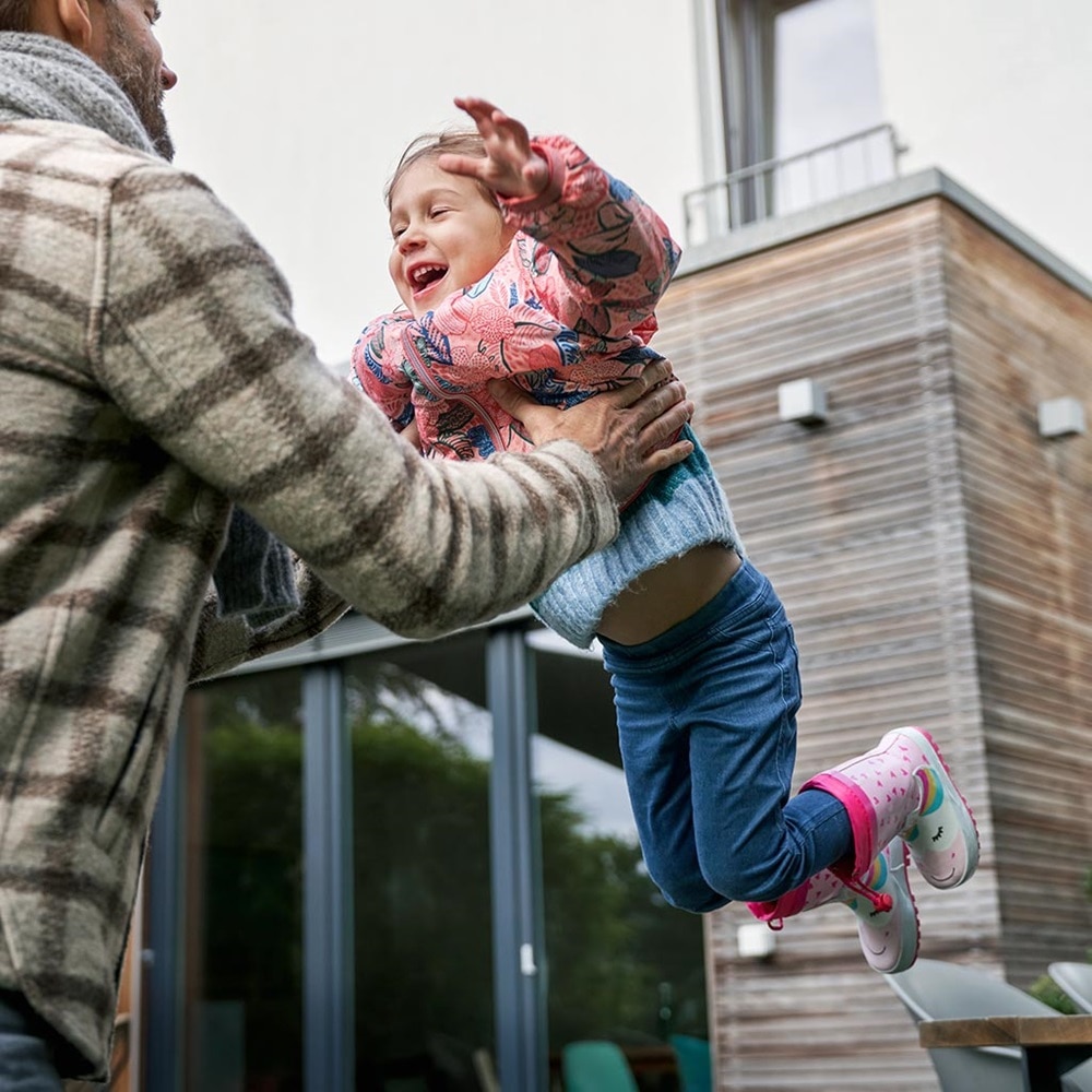 Erwachsene Person hebt ein fröhliches Kind im Freien, mit einem modernen Haus im Hintergrund