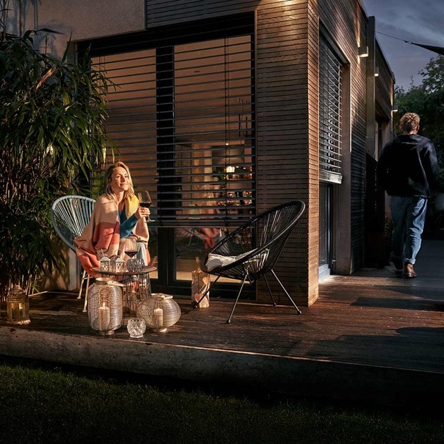 Person sitzt auf Terrassenmöbeln und genießt die Abendstimmung mit Außenbeleuchtung, moderne Gebäudefassade sichtbar