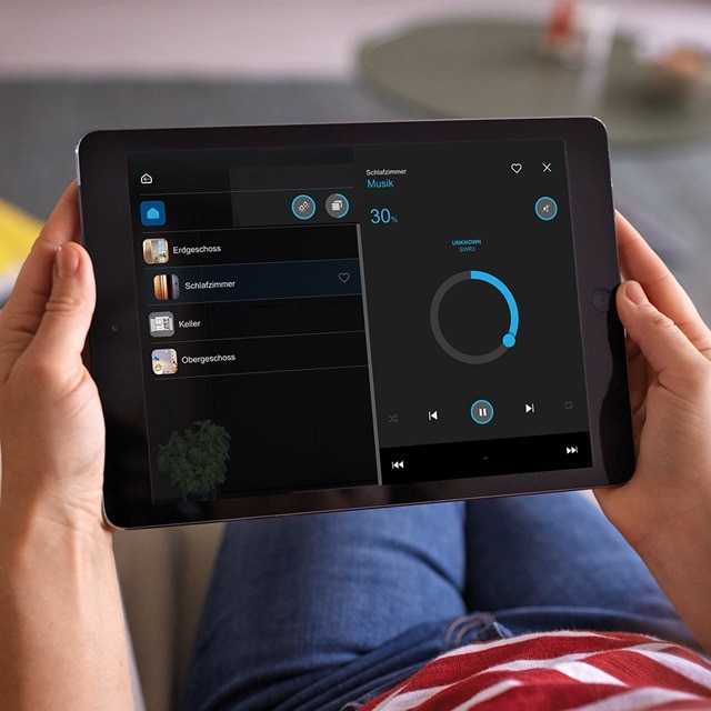 Person hält ein Tablet mit einer Smart-Home-Steuerungs-App-Oberfläche für Beleuchtungs- und Musikanlagen