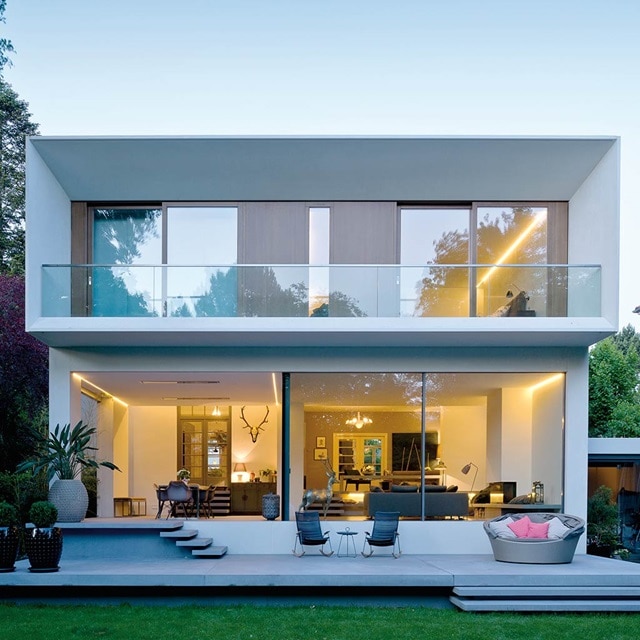 Moderne zweistöckige Hausfassade mit großen Glasfenstern, Außenbeleuchtung und Terrassenmöbeln in der Dämmerung