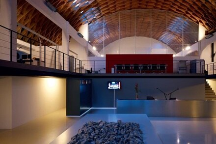Modern loft interior with high ceiling, exposed beams, and lighting fixtures, including visible wall sconces and recessed ceiling lights