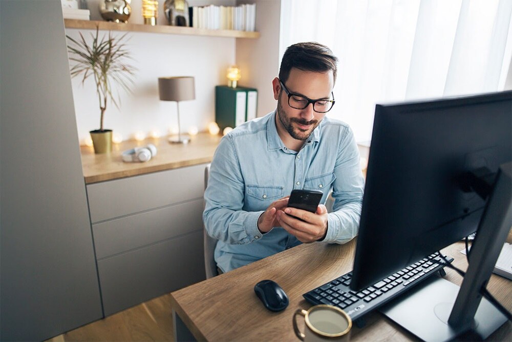Person at a workspace using a smartphone with a computer monitor, keyboard, and mouse on the desk