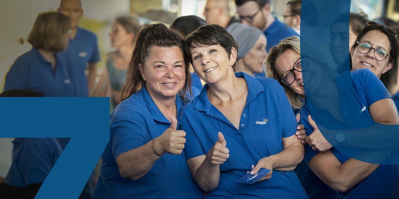 Hager Group employees in blue polo shirts smiling and giving thumbs up at a company event.
