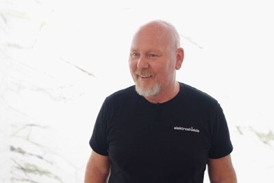 Persona sonriendo con una camiseta negra con texto 'elektroshields' en un espacio interior