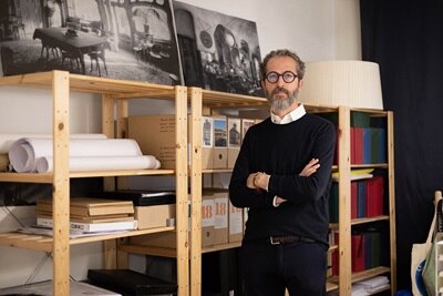 Persona de pie en una oficina con estanterías y varios objetos, incluidos papeles enrollados y libros