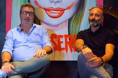 Dos personas sentadas frente a un mural colorido con la palabra 'SER' escrita en él