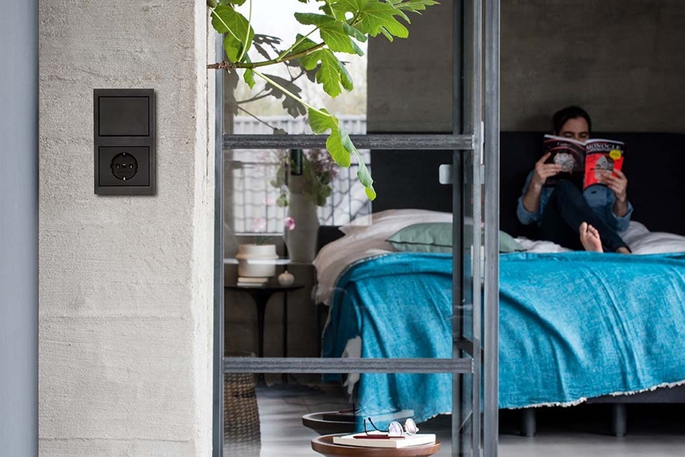 Dormitorio moderno con una persona leyendo en la cama y un enchufe eléctrico de pared negro con interruptor en una pared de hormigón