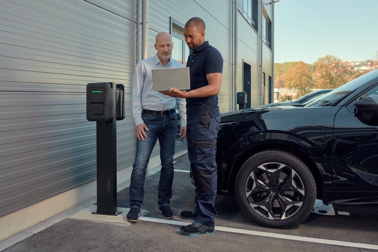 Deux personnes à une borne de recharge pour voitures électriques : l'une avec un ordinateur portable expliquant quelque chose à l'autre, à côté d'une voiture noire et d'un bâtiment moderne avec une façade en métal et de grandes fenêtres en arrière-plan.