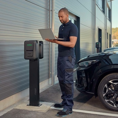 Une personne avec un ordinateur portable à une borne de recharge Hager, à côté d'une voiture électrique noire. La borne est allumée en vert, indiquant qu'elle est opérationnelle. Des bâtiments modernes sont visibles en arrière-plan.