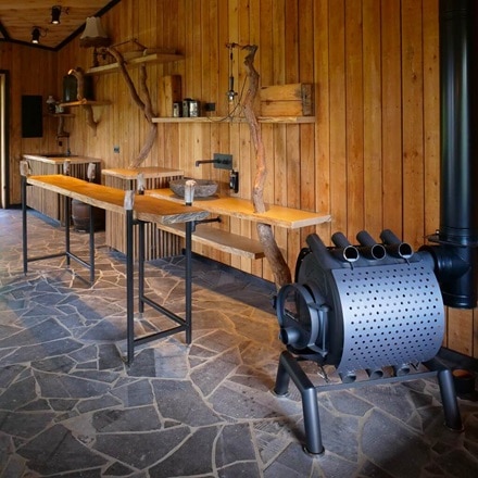 Chambre rustique en bois à l'atmosphère artisanale, meublée de murs en planches de bois, d'un sol en pierres naturelles et d'un poêle à bois noir avec des tuyaux.