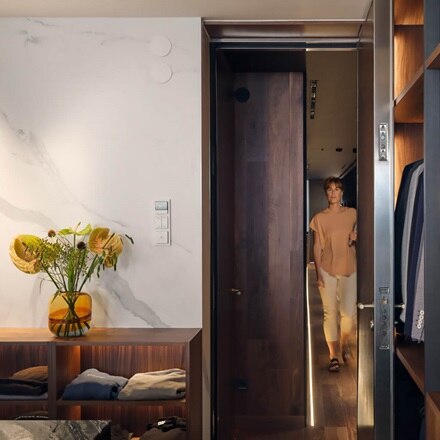 une femme entre dans une loge avec des costumes à droite et des fleurs sur une commode à gauche