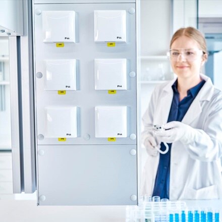 Une femme travaille dans un laboratoire. Elle porte un tablier blanc et des lunettes de sécurité. Au premier plan, six prises électriques, munies de couvercles à charnières, sont accrochées au mur. Des tubes à essai sont posés sur la table.