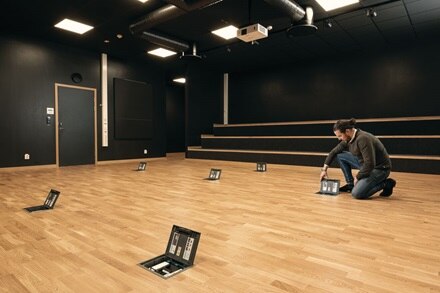 Personne travaillant sur une boîte montée au sol dans une salle de présentation moderne avec tribune, plancher en bois et projecteur au plafond.