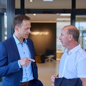 Deux hommes discutent dans un bureau moderne ; l'un en costume bleu, l'autre en chemise blanche avec une veste à la main, contre un mur de verre avec le logo « Hagergroup » en arrière-plan.