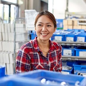 Une personne en chemise à carreaux se tient dans une salle de stockage avec des étagères remplies de bacs bleus et des piles de boîtes blanches, s'intégrant dans un environnement de travail logistique.