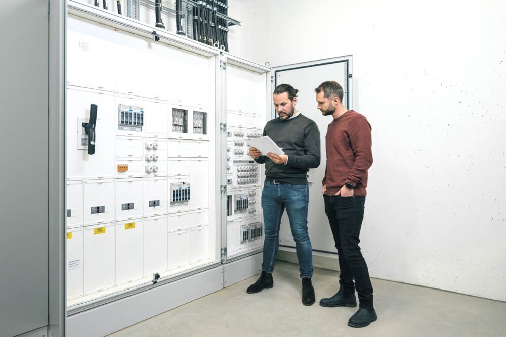 Deux personnes se consultent devant un tableau de distribution électrique avec divers composants dans un environnement de travail technique.