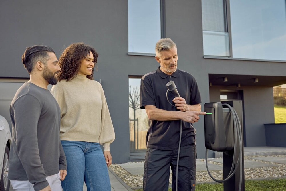 Trois personnes devant une borne de recharge électrique moderne : l'une montre son fonctionnement, tandis que les autres observent avec intérêt. En arrière-plan, un bâtiment moderne.