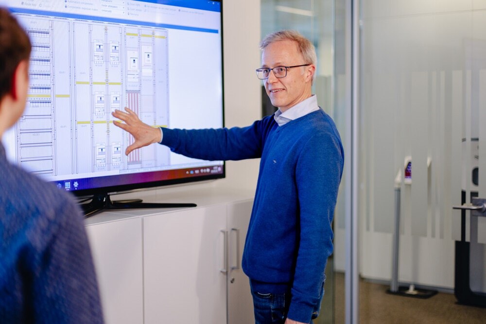 Une personne explique quelque chose à quelqu'un d'autre sur un grand écran posé sur un bureau. Le plan d'un coffret de distribution y est affiché.