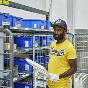 Une personne portant des gants tient un objet blanc dans un entrepôt ordonné avec des bacs bleus étiquetés et des étagères de stockage en métal.
