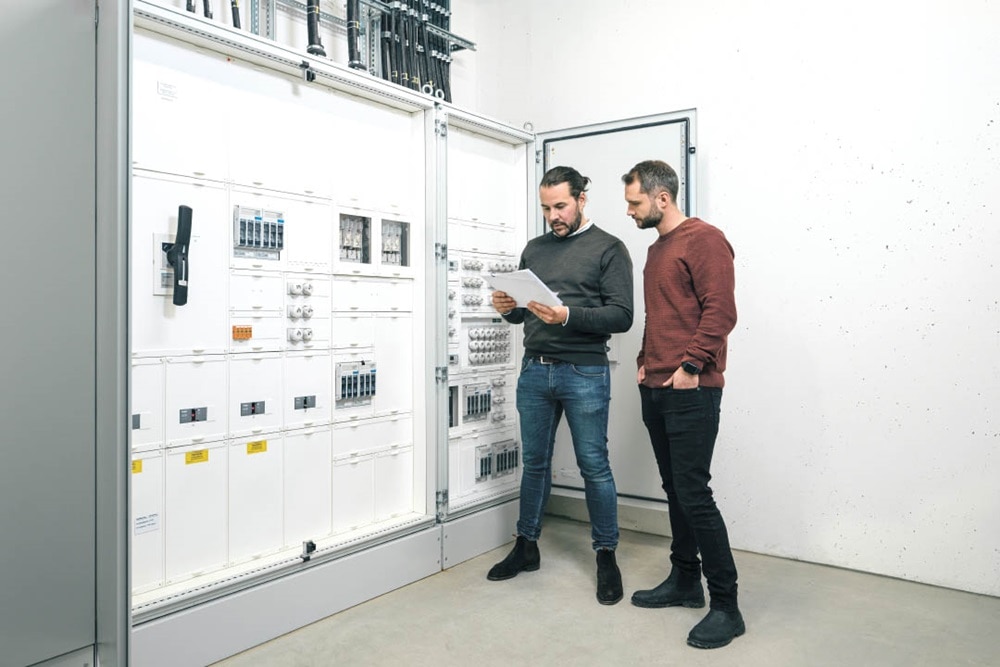 Deux personnes se consultent devant un tableau de distribution électrique avec divers composants dans un environnement de travail technique.