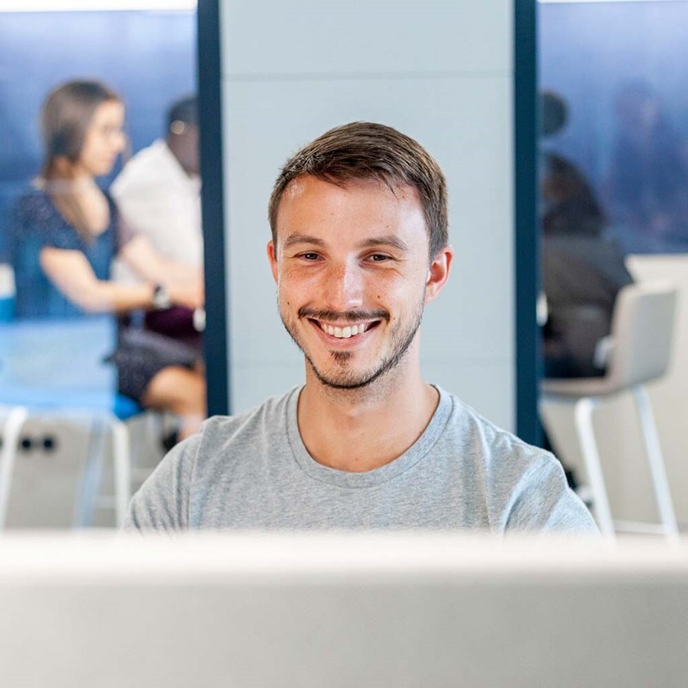 Une personne travaille sur un ordinateur dans un bureau moderne, tandis que deux autres personnes parlent en arrière-plan.