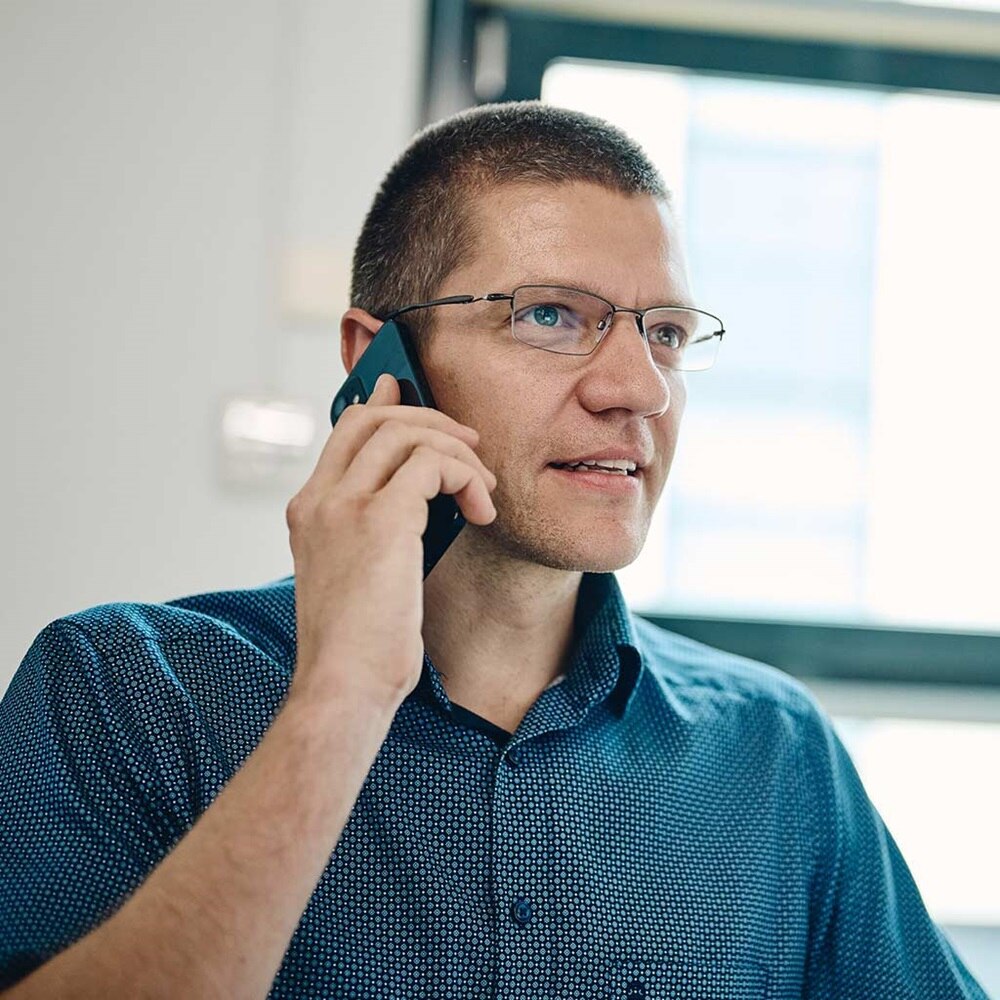 Une personne en chemise bleue tient son smartphone contre son oreille et téléphone. L'arrière-plan est flou, mais laisse entrevoir une fenêtre baignée de lumière naturelle, suggérant un cadre ouvert et professionnel.