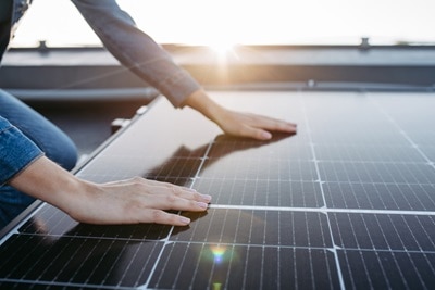 Cette image montre une personne touchant ou installant un panneau solaire avec les deux mains, avec le soleil brillant en arrière-plan.