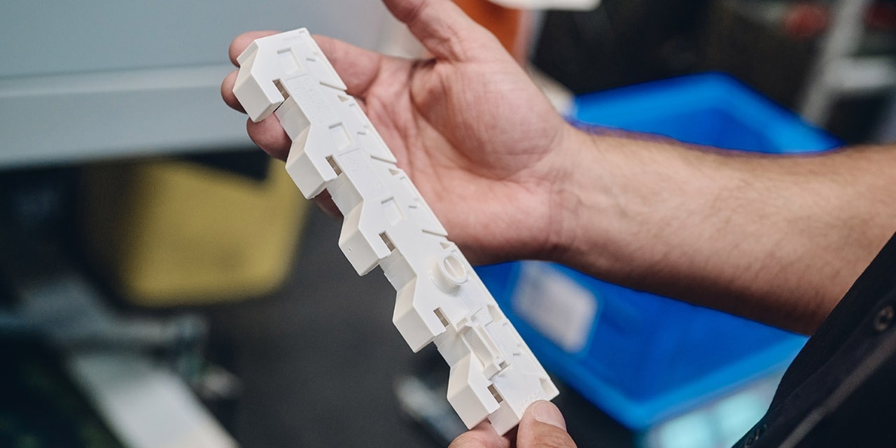 Gros plan de mains tenant une pièce industrielle en plastique blanc dans un environnement d'atelier avec une poubelle bleue en arrière-plan.