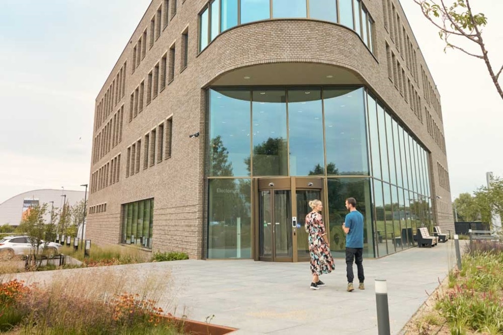 Un immeuble commercial ou de bureaux moderne en briques avec une entrée vitrée. Deux personnes discutent devant l'entrée, dans un cadre urbain verdoyant.