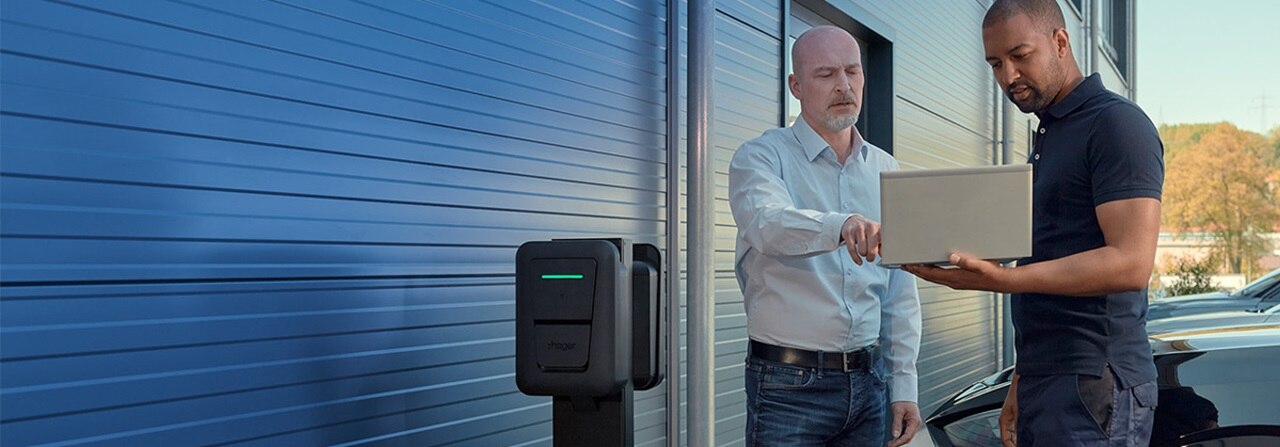 Borne de recharge Witty Pro de Hager installée sur un mur extérieur, avec deux personnes à côté d’un véhicule électrique stationné, vérifiant la configuration et la solution de charge sur un ordinateur portable.