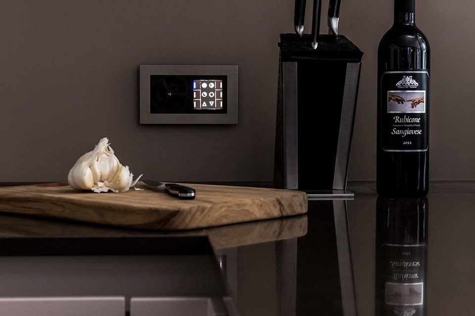 Panneau de contrôle tactile élégant pour domotique fixé au mur de la cuisine à côté d'une bouteille de vin et d'un ensemble de couteaux