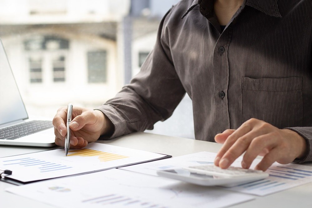 Devant un ordinateur, un homme écrit sur une fiche de la main droite et utilise une calculatrice de la main gauche