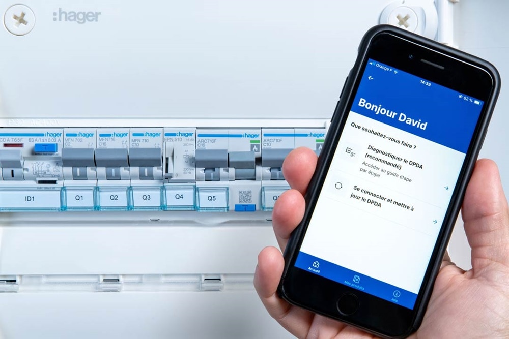 Main tenant un smartphone avec l’app Hager Pilot affichée, devant un tableau équipé de protecteur d'arcs