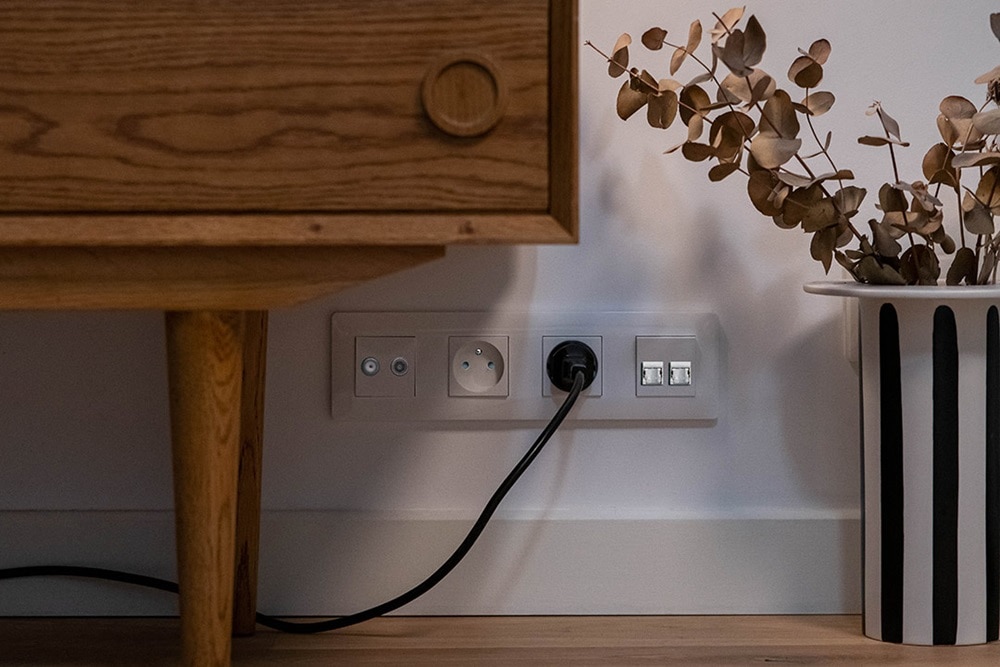 Appareillage essensya sur le bas d'un mur avec une prise TV+FM, deux prises 2P+T et une double prise RJ45, à proximité d'une Commode
