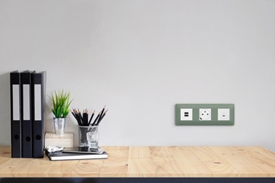 plaques 3 postes essensya botanic de Hager, 2 prises de courant affleurantes et prises USB-C sur un mur devant un plan de travail en bois clair, classeurs noirs et nécessaire de bureau