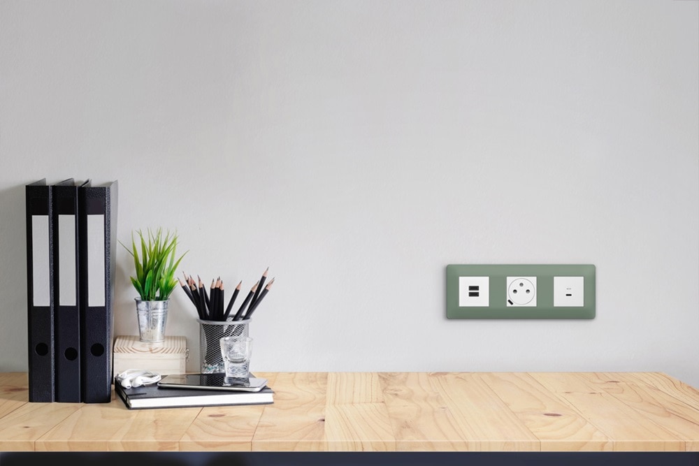 plaques 3 postes essensya botanic de Hager, 2 prises de courant affleurantes et prises USB-C sur un mur devant un plan de travail en bois clair, classeurs noirs et nécessaire de bureau