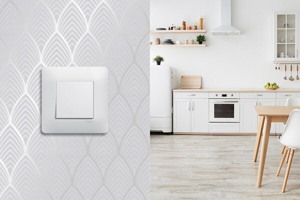 Appareillage mural essensya pure de Hager sur un mur de papier peint blanc aux motifs arts décos laissant apparaitre une cuisine en arrière plan dans les tons de blanc