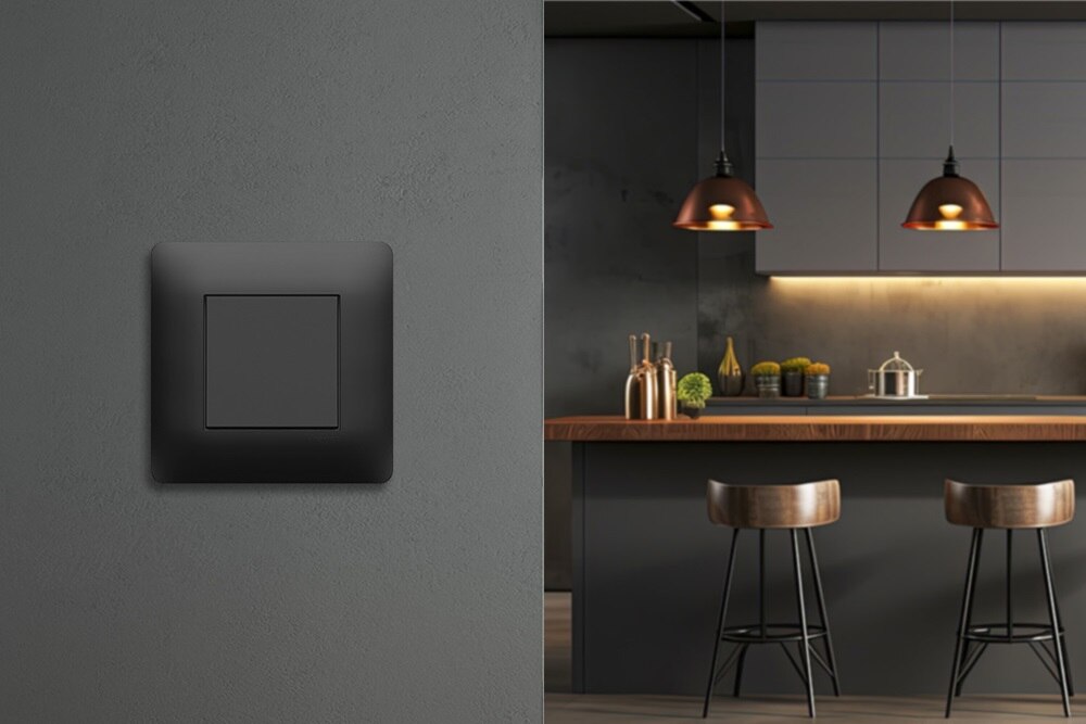 Appareillage mural essensya anthracite de Hager sur un mur gris foncé laissant apparaitre une cuisine en arrière plan dans les tons gris sombre et cuivre