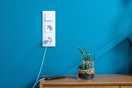 Appareillage mural essensya de Hager, plaque 3 postes, finition pure, fonctions va-et-vient, 2 prises 2P+T alimentant un smartphone posé sur meuble bois à côté d'une plante et mur bleu avec étagère et bougies