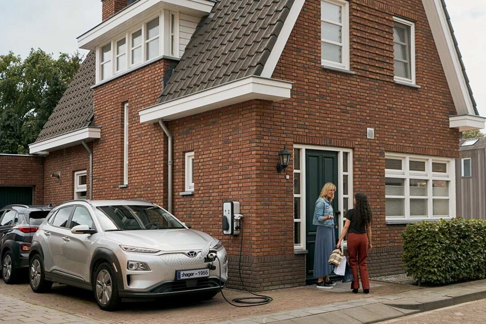 2 femmes discutent sur le perron d'une maison en briques rouges pendant qu'un véhicules gris clair est en charge sur une borne de recharge witty start de Hager fixée sur le mur de côté