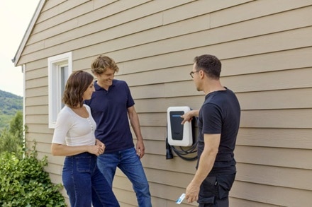 Installateur agréé IRVE qui explique à un couple comment utiliser une borne de recharge witty one version blanche de Hager posée sur le mur de la maison