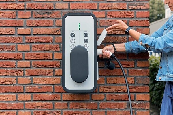 Femme qui branche son câble sur une borne de recharge witty de Hager montée sur le mur en briques rouges de sa maison