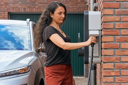 Femme qui branche son câble sur une borne de recharge witty start de Hager montée sur le mur en briques rouges d'une maison