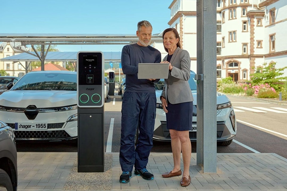 Sur un parking doté de bornes de recharge witty park 2 de Hager, un couple optimise la performance énergétique du parc de bornes de recharge via un ordinateur.