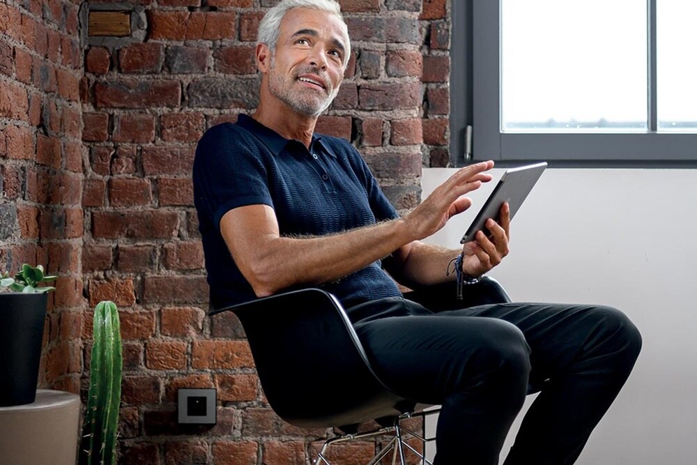 homme aux cheveux blancs qui pilote l'éclairage de son salon depuis sa chaise à bascule à l'aide de domovea de Hager sur son iPad