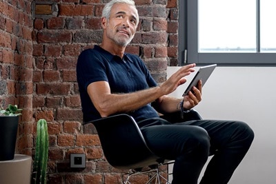 homme aux cheveux blancs qui pilote l'éclairage de son salon depuis sa chaise à bascule à l'aide de domovea de Hager sur son iPad