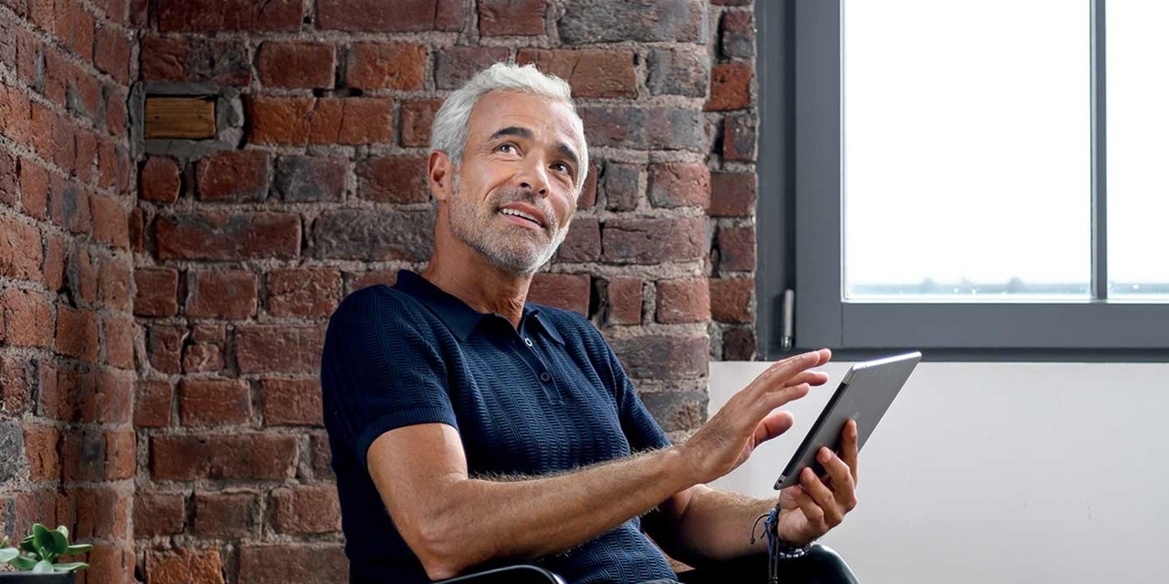 homme aux cheveux blancs qui pilote l'éclairage de son salon depuis sa chaise à bascule à l'aide de domovea de Hager sur son iPad