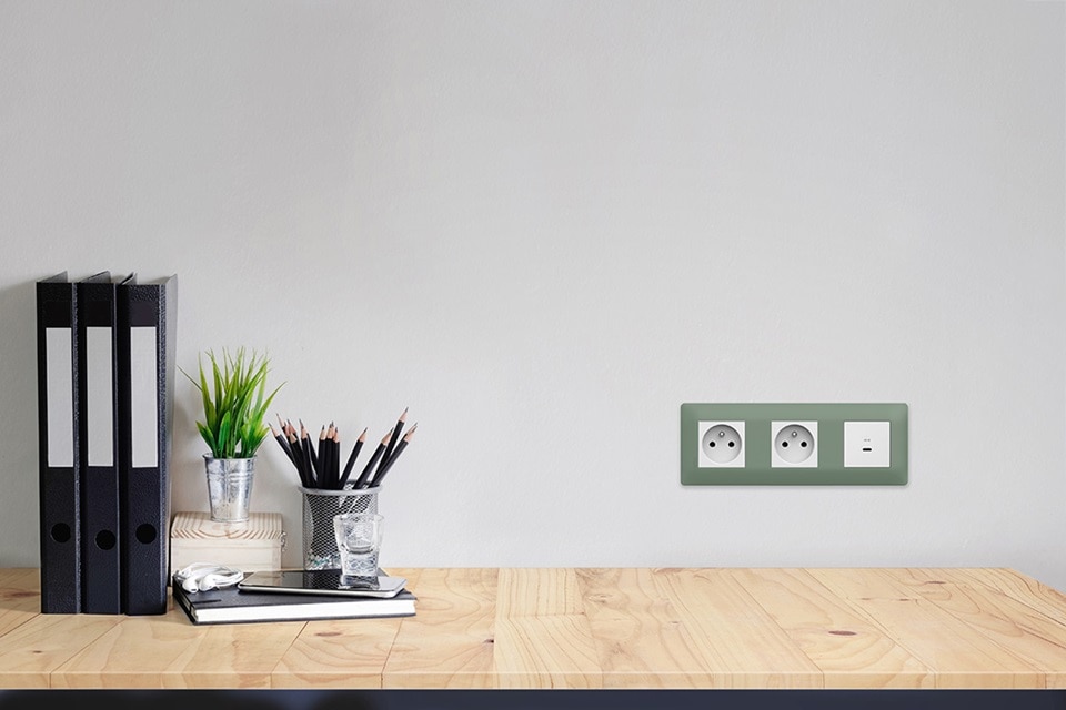 appareillage mural essensya de Hager 3 postes, plaque botanic, 2 prises 2P+T et charge rapide USB-C pure sur le mur blanc d'un bureau en bois clair équipé de classeurs, petite plante verte et pot à crayons.